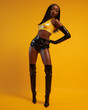 © Pillow Productions - A stunning young African American woman, with long black hair, strikes a pose against a yellow backdrop, dressed in thigh-high black latex boots, gloves, shorts, and a yellow top.