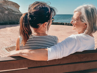 Sticker - Back view of happy couple of elderly women talking sitting on a bench face the sea enjoying sunny day and vacation. Relaxed retirement lifestyle concept