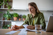 © DimaBerlin - Financial literacy. Focused woman using smartphone calculating expenses, trying to analyze domestic budget, thinking how to pay debt, sitting at kitchen table. Savings, smart spending and consumption.