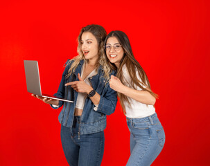 Two excited young best friends reading good news online hold carry laptop pointing screen smiling happy woman clenched fist do winning gesture. Red studio background, copy space.