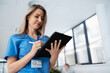 © Halfpoint - Portrait of confident female doctor standing in Hospital corridor. Beautiful nurse wearing blue scrubs, holding clipboard standing in modern private clinic, low angle shot.