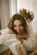 © polinaloves - Portrait of young woman in white knitted dress, she is sitting in a chair in a cozy room.
