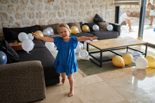 Happy Girl Standing With Arms Outstretched With Birthday Balloons In Living Room At Villa