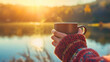 A woman wearing a sweater, holding a mug or a cup in her hands, standing outdoors, looking at the sunny lake and forest landscape in the morning. Tourist or vacation hot coffee drink, weekend beverage