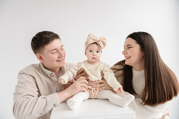 Dad, mom holds in hands 6 month old little kid. Father, mother hugging daughter. Happy family hugs cute baby on a holiday closeup. Daddy, mommy embracing girl with bow in hair isolated on white wall.