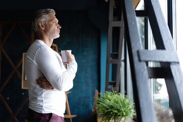 Wall Mural - Businessman taking a coffee break standing leaning against a windowsill turning to stare out with a thoughtful expression.