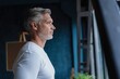 © opolja - Attractive mature man leaning on the sill of a large bright window with copy space looking away.