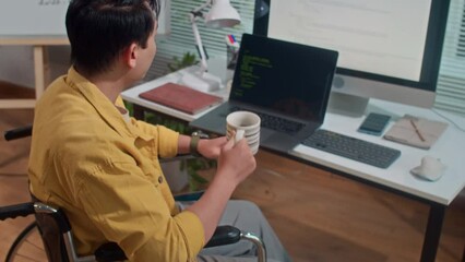 Tilt up shot of young male developer in wheelchair drinking coffee while testing code on computer and laptop at office