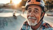 An elderly man with a gray beard and mustache wearing a red helmet smiling at the camera sitting on a skateboard ramp with a blurred background of a skate park.