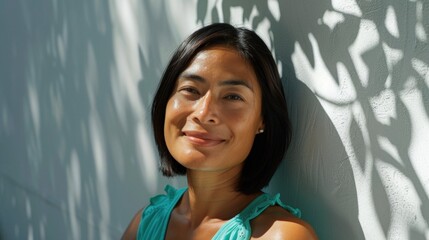 Poster - Smiling woman with dark hair and a light blue top leaning against a wall with sunlight and shadows from leaves.