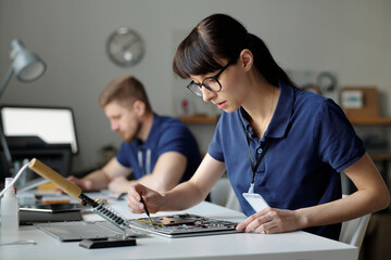 Pretty technical service worker fixing tiny details of disassembled laptop while sitting by workplace in front of camera against colleague