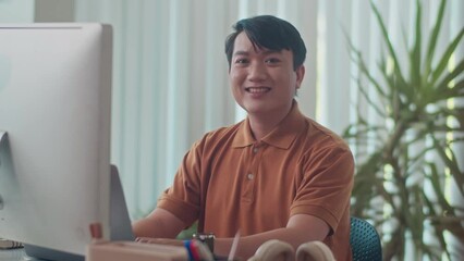 Medium portrait of cheerful office employee of Asian ethnicity sitting in front of monitor and looking at camera