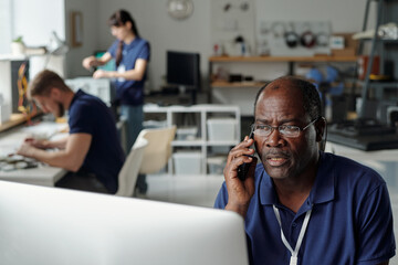 Aufkleber - Mature male technician of repair service sitting by workplace in front of computer screen and talking to client on mobile phone