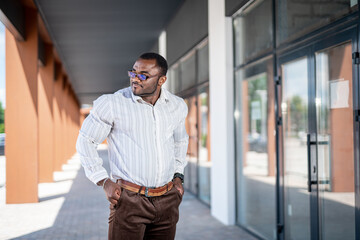 Wall Mural - Stylish  man in white shirt and brown pants stands on the street near the building and looks away. 