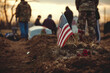 American flag by fresh grave at military funeral, with soldiers and mourners in the background.