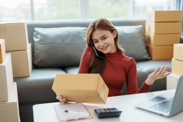 Young woman entrepreneur with a package while on a phone call at home. Online business and customer service concept.