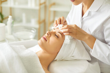 Wall Mural - Close-up photo pf professional cosmetologist or beautician doctor hands making anti-age procedures cleaning with napkins facial mask of young pretty woman client in beauty spa clinic.