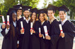 Group of happy cheerful diverse multiethnic college or university students and friends wearing black graduation caps and gowns, with diplomas in hands, standing together, looking at camera and smiling