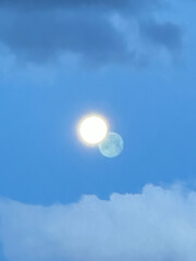 Image taken over Cody, Wyoming, February 25, 2024, shows a bright moon against a blue sky with a shadow image in the distant clouds. Double moon with yellow circle looks like two.. 