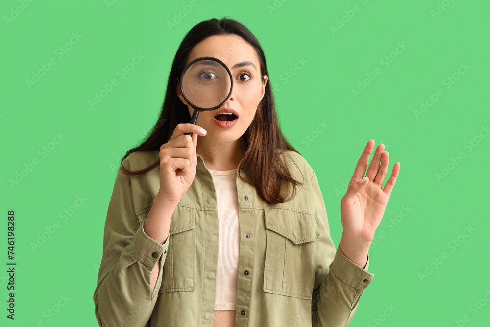 Shocked young woman with magnifier on green background
