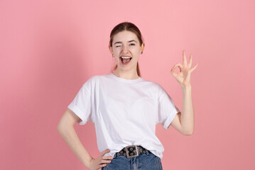 Wall Mural - Attractive caucasian young brunette woman in casual white t-shirt winks and shows okay gesture isolated on pink studio background.