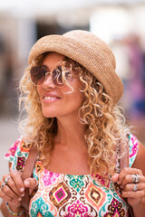 Sticker - Happy tourist woman with blonde long curly hair and hat looking around and walking alone with backpack enjoying outdoor leisure activity and holiday vacation time. People defocused in background.