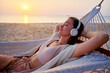 © Goffkein - Young blissful calm woman wearing wireless headphones with closed eyes and hands behind head lie in a hammock and enjoy relaxing music