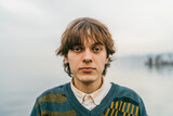 Fototapeta  - Young male with contemplative gaze, wearing sweater, against blurred lakeside backdrop.