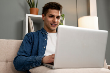 Young man relaxing on the sofa with a laptop at the living room. Concentrated smiling male sitting in home working from home with computer.