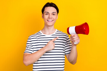 Poster - Photo of positive cheerful guy with stylish hairdo dressed striped t-shirt indicating at loudspeaker isolated on yellow color background