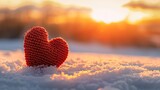 Fototapeta Niebo - A handmade red knitted heart rests on fresh snow, backlit by a golden sunset, symbolizing warmth and love in winter.