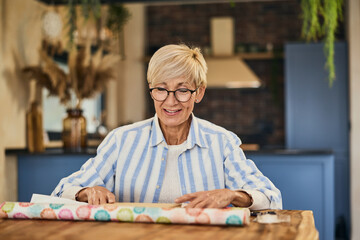 Wall Mural - A happy senior woman using a wrapping paper and packing a gift for someone, at home.