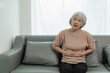 © ArLawKa - An elderly woman has severe abdominal pain. Sitting alone on the sofa while having dinner at home, elderly health care concept. Illness. Health insurance.