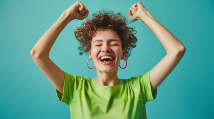 Wall Mural - joyful person with curly hair, raising their arms in excitement, wearing a green shirt and red earrings, with a wide, laughing smile against a blue background.