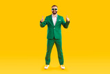 Fototapeta Natura - Full body length shot of happy confident handsome bearded young man in trendy funky green suit and sun glasses standing on yellow studio background, smiling and giving thumbs up with both hands