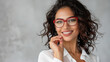 A confident woman holding a pair of red glasses, her joyful expression and elegant white shirt complemented by her stylish curly hair, against a neutral background.