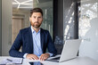 © Tetiana - Portrait of a successful and confident young man lawyer in a business suit sitting at an office desk, working with a laptop and documents, looking seriously at the camera