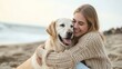 A young woman in a sweater hugs a golden retriever dog sitting on the sand near the sea