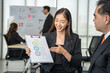 © Bavorndej - Cheerful Asian businesswoman presenting a clipboard with ESG (Environmental, Social, and Governance) concepts to a male colleague.