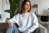 Fototapeta  - Mockup. Young woman wearing blank white crewneck sweatshirt. Young female sitting on sofa in modern living room. Mock up template for sweatshirt design, print area for logo or design