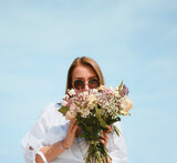 Fototapeta Desenie - Joyful Young Woman Holding a Bouquet of Flowers on a Beach with Blue Sky Background