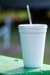 A large white nonrecyclable to-go drink cup with a plastic lid and straw on a vibrant green patio table. The polystyrene or Styrofoam insulated cup is filled with a cold soda pop drink. 
