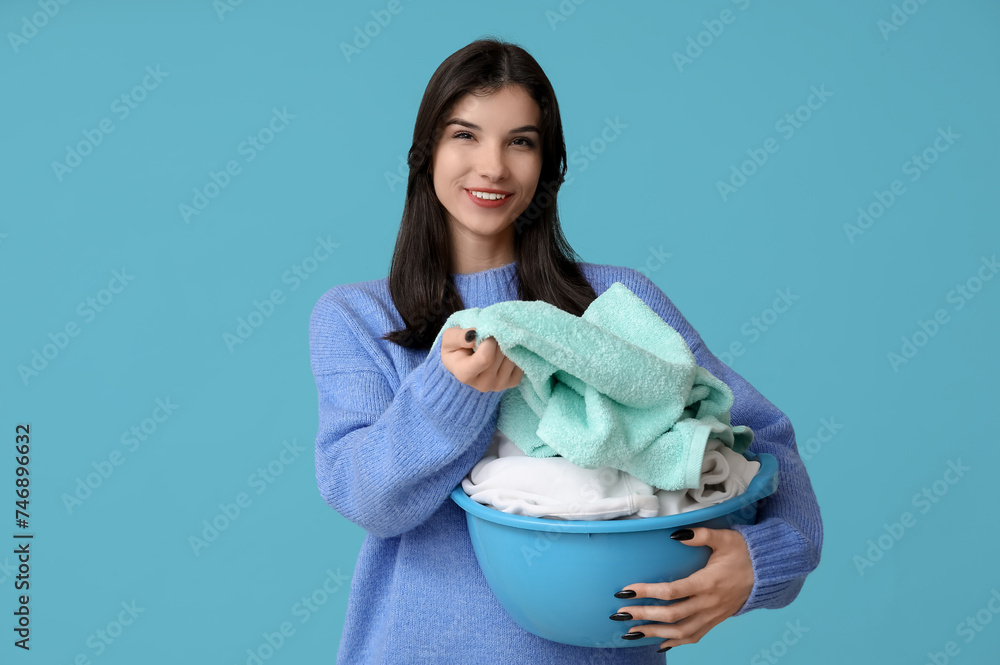 Happy young woman holding basket with clean laundry on blue background