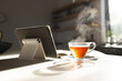Steam rises from a hot cup of tea next to a tablet on a sunlit table