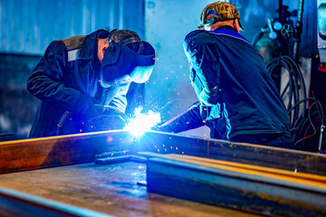 A welder wearing a protective mask for metal welding and protective gloves performs welding work at a metal structures plant. Welding a steel industrial beam. Sparks, hot flame from welding.