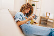 © SHOTPRIME STUDIO - Couch Chat: Young Woman Holding a Smartphone, Smiling at the Camera on a Cozy Sofa in her Home