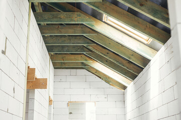 Unfinished attic wooden roof framing with vapor barrier, dormer and windows. View on timber rafters and beams on aerated concrete blocks in mansard. New modern farmhouse construction