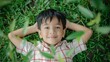 © iuricazac - A young boy with black hair wearing a red and white checkered shirt lying on green grass with his arms behind his head smiling and looking at the camera.