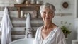 © iuricazac - A woman with short gray hair wearing a white blouse standing in a bathroom with a wooden wall and a potted plant in the background.