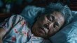An elderly woman with gray hair peacefully sleeping on her side with her eyes closed wearing a floral patterned shirt resting on a blue pillow in a dimly lit room with a blurred background.